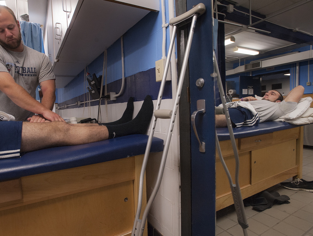 University of Maine trainer Ben Randall works on defensive end Kurt Massey, reflected at right. Massey says the school should pay his medical bills, but he’s not bitter and he’s not giving up: “Football’s one of those sports where you put your whole heart into it or you put nothing into it.”