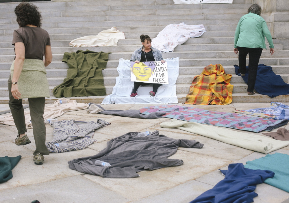 Joanie Klayman, left, and Caroline Fernandes, right, both of Preble Street, place blankets on the steps of Portland City Hall last Tuesday, while Mary Jo Skofield from Homeless Voices for Justice holds a sign in protest of potential budget cuts to homeless overflow shelters in Portland.