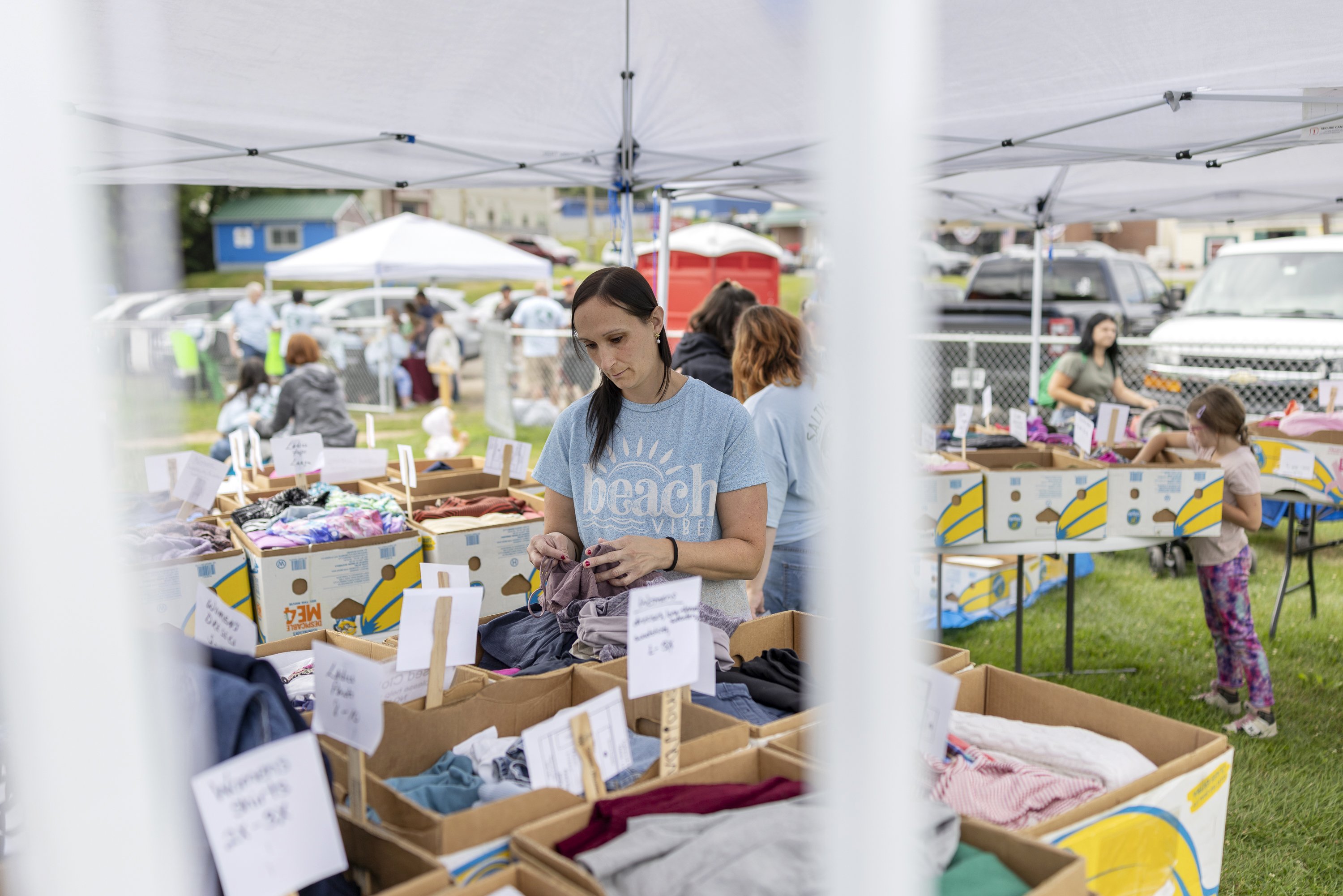 A woman stands outside under a tent. Surrounding her are many boxes full of kids' clothing.